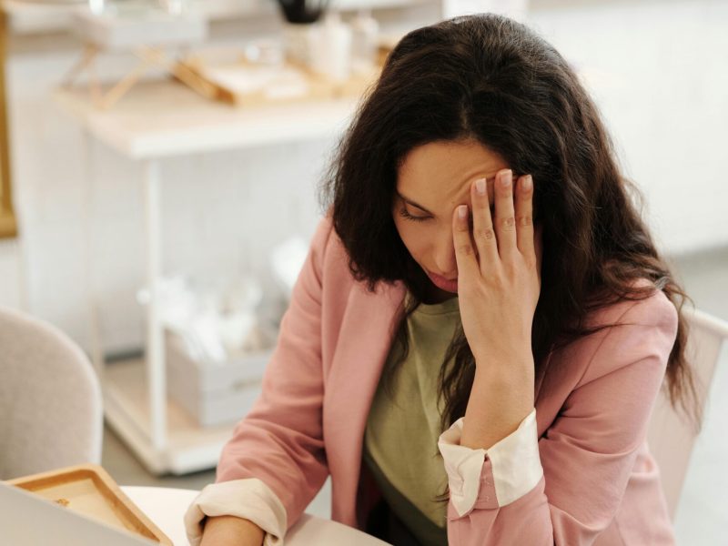 Woman holding head in hands while working on a laptop indoors, seemingly stressed or unwell.
